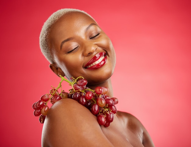 Soins de la peau sourire et raisins avec une femme noire modèle en studio sur fond rouge pour la santé ou la nutrition Beauté heureuse et fruit avec une jeune femme posant pour un régime de bien-être ou de désintoxication