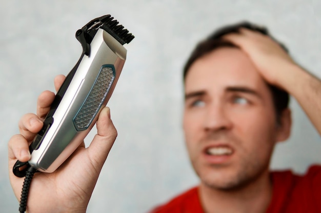 Soins personnels dans les conditions de quarantaine mondiale et de salons de coiffure et de beauté fermés Bel homme se coupant les cheveux avec une tondeuse et regarde dans le rasoir électrique de la tondeuse