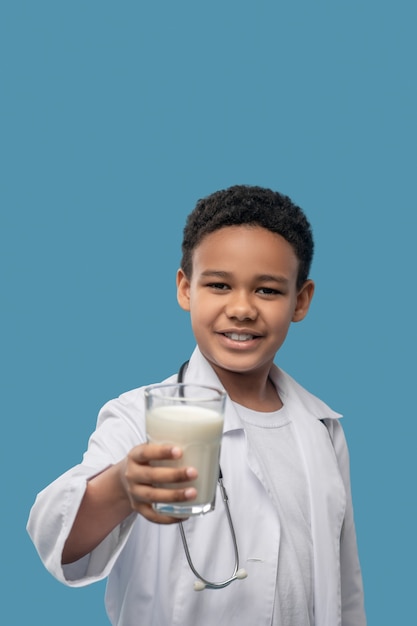 Soins de santé. Heureux garçon afro-américain en blouse blanche médicale et stéthoscope offrant un verre de lait en photo de studio