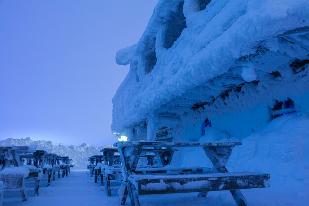 Soirée d'hiver. Café en plein air vide au sommet de la piste de ski. Maison couverte de neige. Tables et bancs