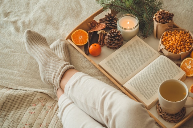 Soirée d'hiver douillette, chaussettes en laine chaudes. La femme est allongée les pieds sur une couverture blanche et un livre de lecture. Scène de loisirs confortable. Le texte du livre est illisible. Femme de détente à la maison. Style de vie confortable.