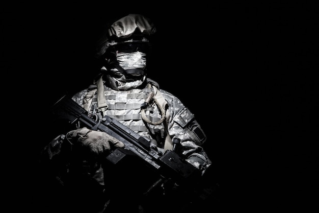 Soldat des Forces armées des États-Unis en tenue de combat avec des lunettes noires et un masque sur le visage, l'arme automatique de l'escouade armée émerge de l'obscurité. Menace militaire, mission furtive secrète, combattant de guerre hybride