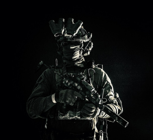 Photo soldat des forces d'opérations spéciales de l'armée en masque et uniforme de combat casque équipé d'un dispositif de vision nocturne mitraillette armée avec silencieux regardant de côté portrait de studio discret sur fond noir