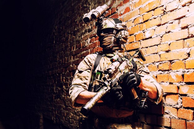 Photo soldat des forces d'opérations spéciales combattant de l'équipe d'assaut contre le terrorisme avec dispositif de vision nocturne sur casque et fusil de service mur de briques de tir intérieur discret