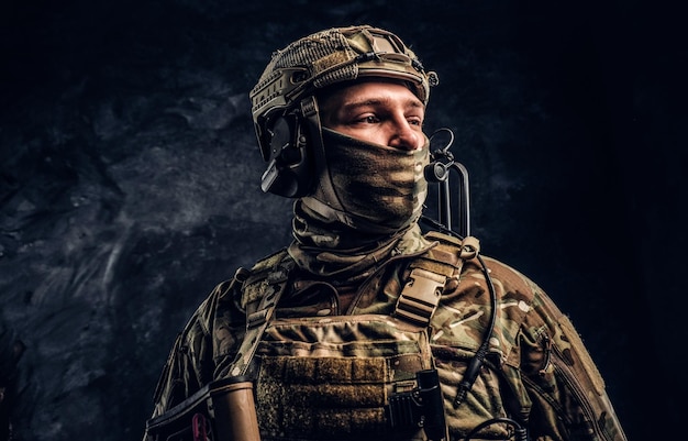 Soldat des forces spéciales modernes en uniforme de camouflage regardant de côté. Photo de studio contre un mur texturé sombre.