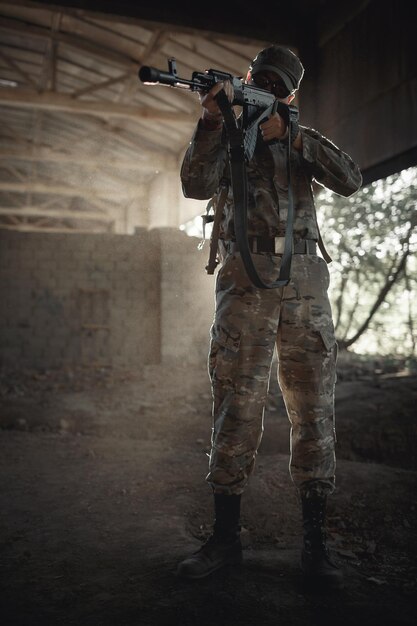 Un soldat intrépide dans un uniforme de protection camouflage dans des lunettes noires tactiques et dans une casquette avec une mitrailleuse dans ses mains se tient à l'intérieur d'une grande maison autorisée