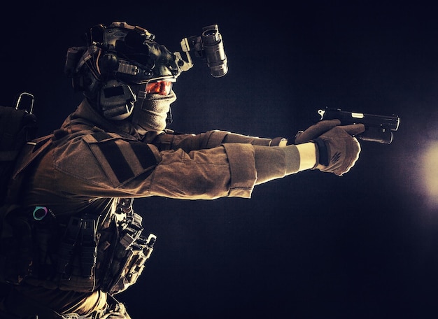 Photo soldat moderne combattant de l'escouade anti-terroriste dans un casque uniforme de combat et un casque radio tactique visant un pistolet de service dans l'obscurité tirant avec un portrait de studio d'arme de poing sur fond sombre