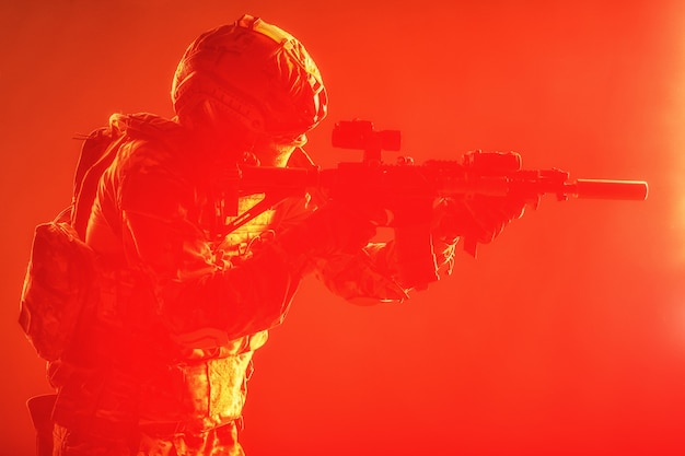 Photo soldat moderne des forces spéciales de l'armée, combattant de l'escouade antiterroriste de la police en uniforme de combat, casque avec dispositif de vision nocturne visant un fusil d'assaut à canon court, tournage en studio discret avec rétroéclairage rouge