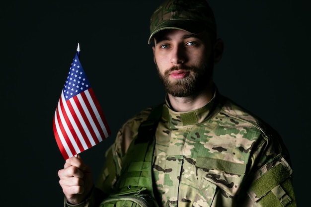 Un soldat sérieux en uniforme militaire tient le drapeau américain regarde la caméra confiant et sérieux en studio sur fond sombre