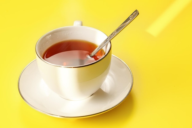 Le soleil du matin brille sur une tasse en porcelaine blanche avec du thé noir chaud, juste infusé, une cuillère en argent, sur un tableau jaune