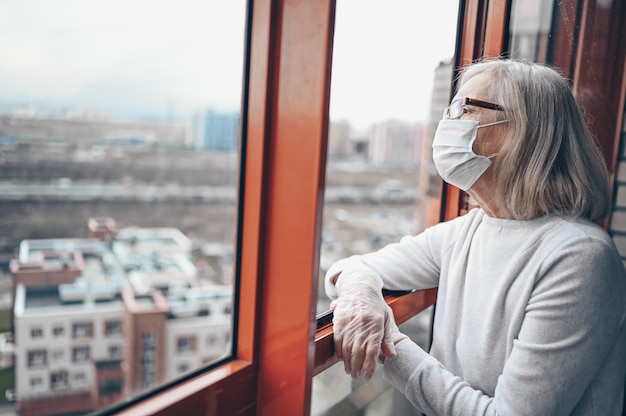 Solitaire femme âgée âgée triste dans un masque médical et des gants en regardant la fenêtre reste l'isolement à la maison pour l'auto-quarantaine. Concept de quarantaine à domicile, prévention COVID-19, situation de flambée de coronavirus