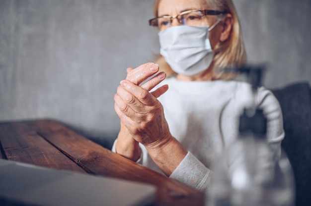 Solitaire triste femme âgée âgée face à un masque médical utilisant un désinfectant liquide antibactérien pour les mains avec un ordinateur portable à la maison quarantaine d'isolement pendant la pandémie de coronavirus COVID19. Rester à la maison