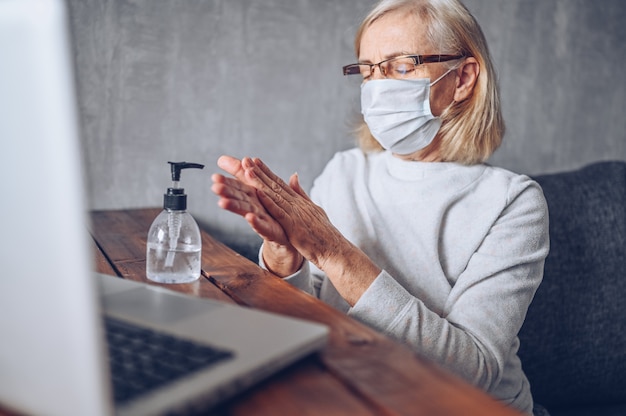 Solitaire triste femme âgée âgée face à un masque médical utilisant un désinfectant liquide antibactérien pour les mains avec un ordinateur portable à la maison quarantaine d'isolement pendant la pandémie de coronavirus COVID19. Rester à la maison
