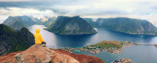 Solitude avec la nature Vue aérienne panoramique sur le fjord et le village de pêcheurs Homme touriste assis sur une falaise de roche Beau paysage de montagne Nature Norvège Îles Lofoten