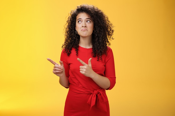 Photo sombre bouleversée et triste jolie fille idiote aux cheveux bouclés en robe rouge pinçant les lèvres dans un sourire malheureux fronçant les sourcils de regret, ratant une chance ou une offre pointant vers le coin supérieur gauche sur fond jaune.