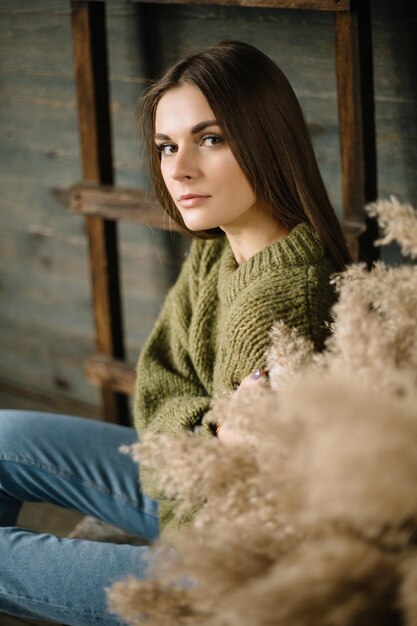 Sombre fille brune dans un pull chaud en jeans et baskets posant en studio avec une échelle de fond en bois à la lumière du jour près du mur