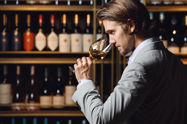 Photo sommelier appréciant le vin blanc du verre en l'odeur debout dans la cave contre l'étagère remplie