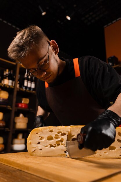 Un sommelier au fromage dans un magasin d'alimentation coupe un maasdam naturel vieilli et vérifie la qualité Bel homme barbu travaillant dans un magasin d'alimentation