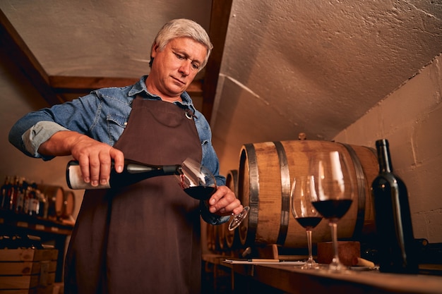 Sommelier professionnel concentré travaillant dans la cave à vin avec des tonneaux en arrière-plan et versant du vin dans un verre