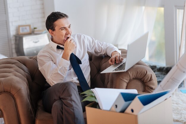 Somnolent. Homme élégant fatigué ennuyé bâillant tout en regardant l'écran de son ordinateur portable moderne et se sentir somnolent alors qu'une boîte d'objets personnels debout à ses côtés