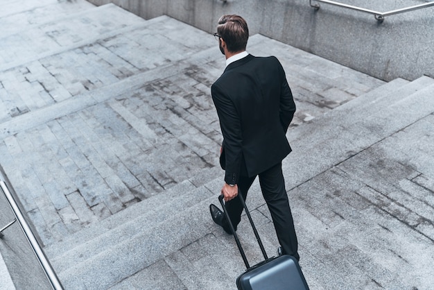 Son travail ici est terminé. Vue arrière supérieure du jeune homme en costume complet tirant des bagages