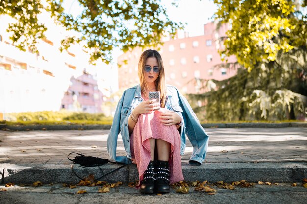 Songeuse jeune femme avec des lunettes de soleil assis sur le trottoir en regardant un téléphone mobile en attente de quelqu'un dans le parc. Émotion humaine expression du visage, sentiment, langage corporel de réaction. Concept émotionnel.