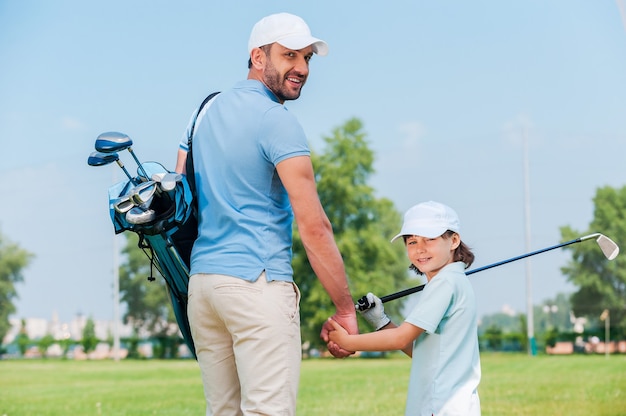 Ce sont les grands fans de golf. Heureux jeune homme et son fils se tenant la main et regardant la caméra en marchant sur le terrain de golf