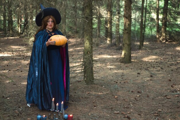 Une sorcière aux cheveux bruns dans un chapeau et un manteau bleu dans la forêt avec une énorme citrouille dans ses mains brûlant des bougies sur le sol