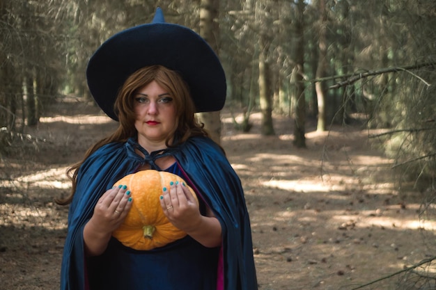 Une sorcière aux cheveux bruns dans un chapeau pointu et un manteau bleu dans la forêt avec une énorme citrouille dans ses mains