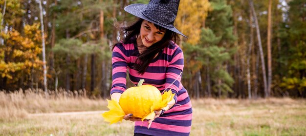 Sorcière souriante d'Halloween avec une citrouille dans une forêt d'automne. bannière pour la fête de Palloween