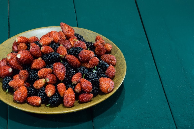 Une soucoupe avec des fraises et des mûres se dresse sur une table verte en bois à gauche