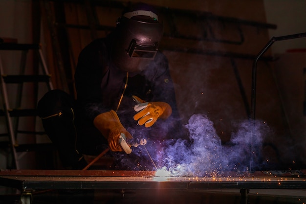 Soudeur assis dans l'acier de soudage en usine pour le rendre plus fort. Le travail produit des étincelles brillantes avec de la fumée, l'effet de la lumière et de la fumée lui donne une belle lumière violette.