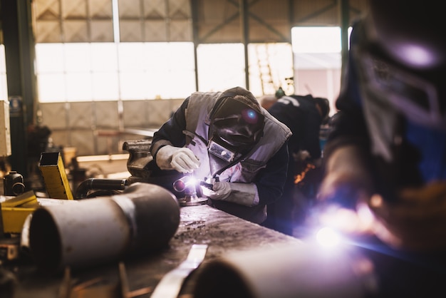 Soudeur en uniforme de protection et masque de soudure de tuyau métallique sur la table industrielle pendant que des étincelles volent.