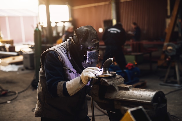 Soudeur en uniforme de protection et masque de soudure de tuyau métallique sur la table industrielle pendant que des étincelles volent.