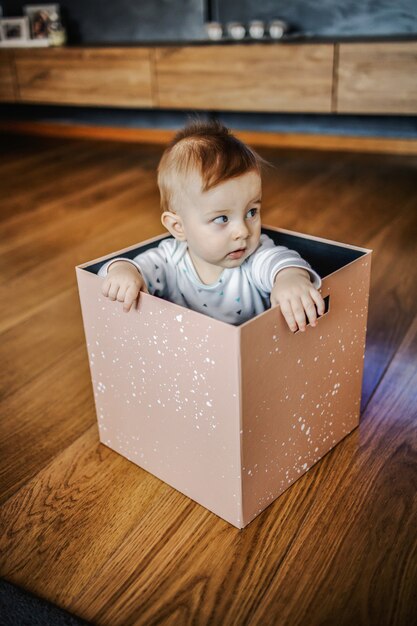 Soupçonneux adorable petit garçon blond assis dans la boîte et regardant ailleurs. Intérieur de la maison.