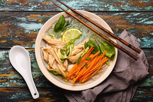 Soupe asiatique avec nouilles de riz, poulet et légumes dans un bol en céramique servi avec cuillère et baguettes sur fond de bois rustique d'en haut, cuisine chinoise ou thaïlandaise