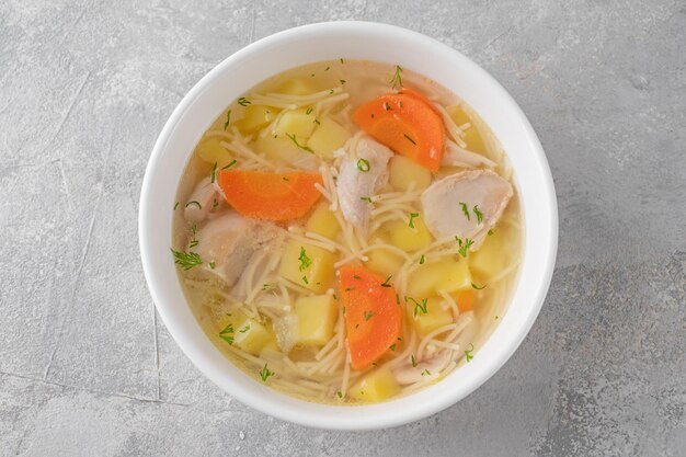 Soupe au poulet avec nouilles et légumes dans un bol blanc sur fond de béton gris