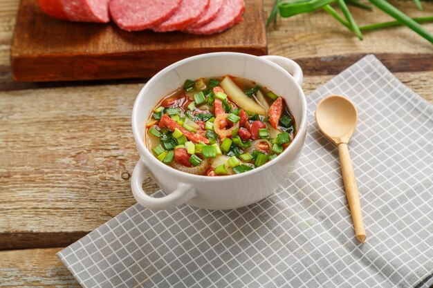 Soupe aux haricots juive avec viandes fumées dans une soupière sur une serviette grise à côté d'une saucisse et d'une cuillère en bois