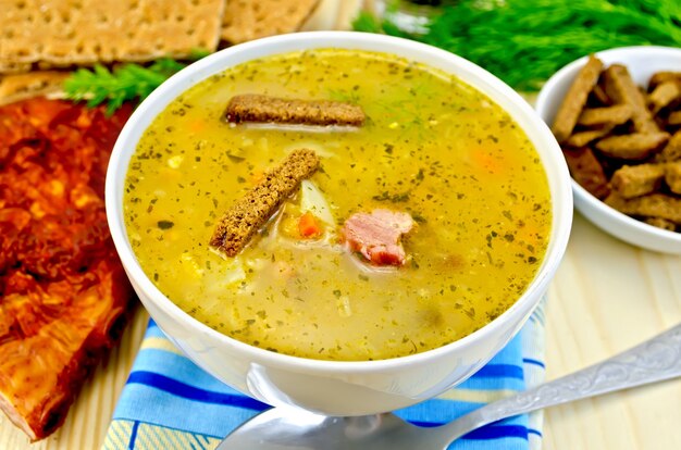 Soupe aux pois avec croûtons de seigle dans un bol et sur une assiette, cuillère, poivre, pain, chiffon, bacon, aneth contre une planche de bois