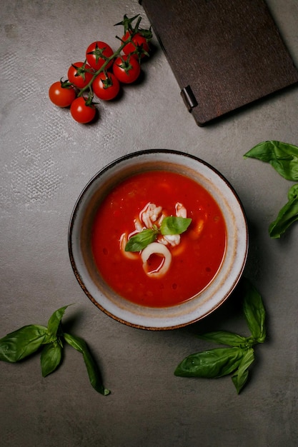 soupe aux tomates avec calamars dans une assiette sur fond gris, à côté d'un menu en bois et de tomates, vue de dessus