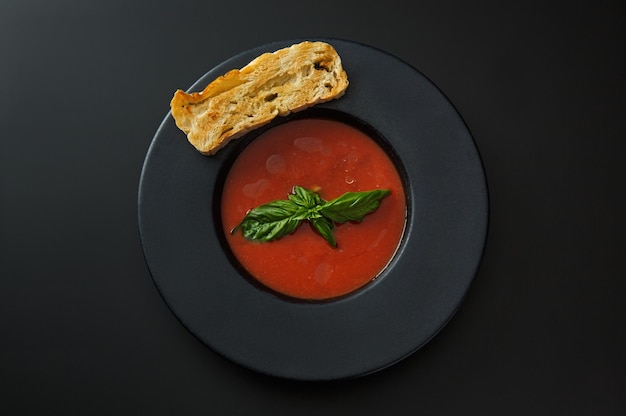 Soupe aux tomates avec un toast et basilic dans une assiette noire isolée sur fond noir