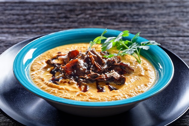 Soupe à la citrouille de purée de citrouille rôtie et bouillon de champignons sauvages, garnie de graines de sésame, plat réconfort végétalien, servi sur une assiette bleue avec cuillère et ail sur fond de bois sombre, vue de dessus