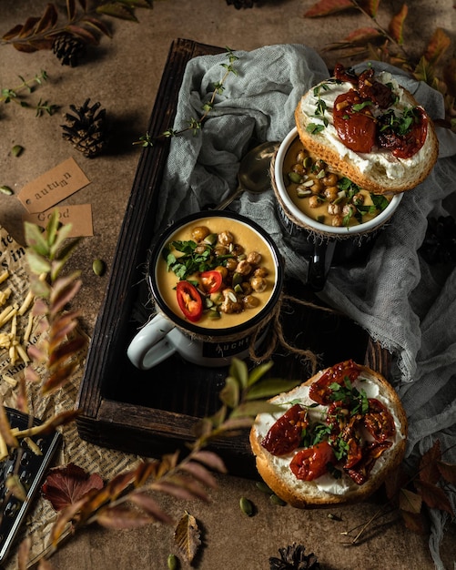 Soupe à la crème de pois chiches dans une tasse dans un plateau en bois Il y a un sandwich au fromage et aux tomates séchées sur la tasse Vue de dessus