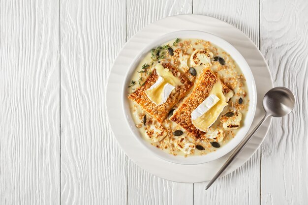 Soupe crémeuse de chou-fleur végétalien garnie de toasts au camembert fondu dans un bol blanc sur une table en bois, vue horizontale au-dessus, gros plan, plat, cuisine française