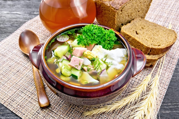 Soupe froide okroshka de saucisses, pommes de terre, œufs, radis, concombre, légumes verts et boisson de kvas dans un bol en argile, pain sur serviette sur fond de planche de bois foncé