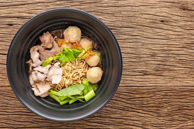 Soupe de nouilles instantanées avec boulettes de viande de boeuf foie de porc eau épinards gloire du matin céleri ail frit et herbes dans un bol en céramique noire sur fond de texture de bois rustique vue de dessus à plat