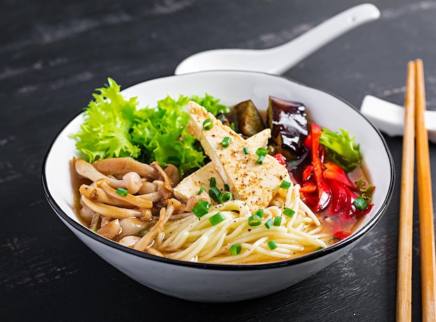 Soupe de nouilles végétaliennes asiatiques avec tofu, fromage, champignons shimeji et aubergines frites dans un bol sur un fond sombre