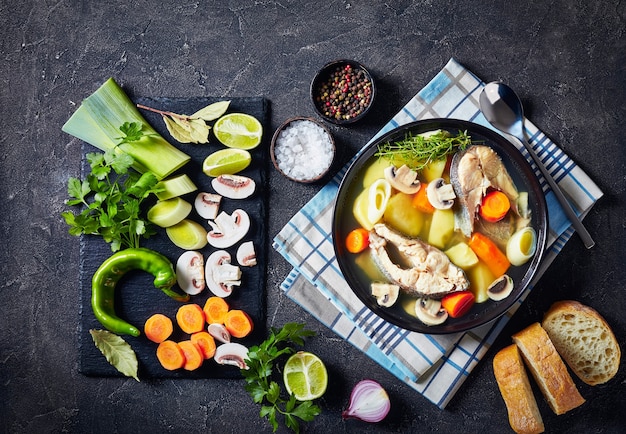 Soupe de poisson à la truite avec légumes et champignons servie dans un bol noir sur une table en béton avec des ingrédients sur une ardoise, cuillère en argent sur une serviette, vue horizontale d'en haut, espace libre, plat