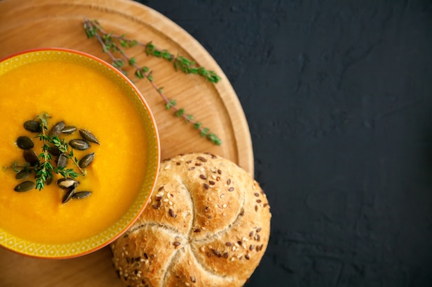 Soupe de potiron maison en gros plan avec du fromage, servi avec des graines, des herbes et du pain, sur fond noir.