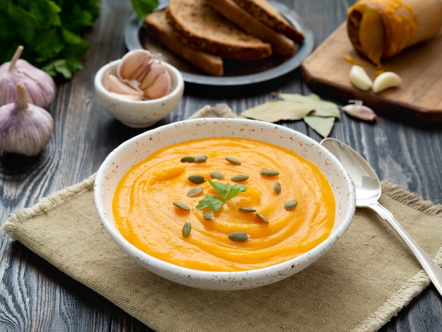 Soupe de potiron en purée dans une assiette blanche, graines de citrouille, pain, ail sur fond en bois foncé